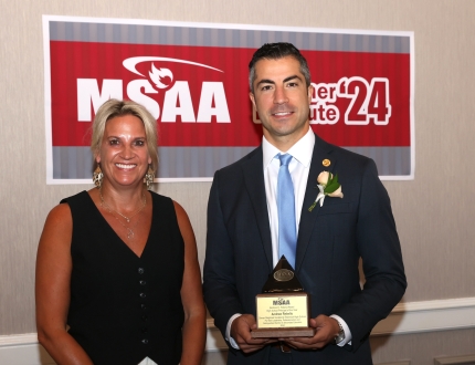 two people stand in front of a msaa sign, one is holding an award.