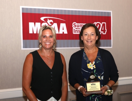 two people stand in front of a msaa sign, one is holding an award.