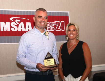 two people stand in front of a msaa sign, one is holding an award.